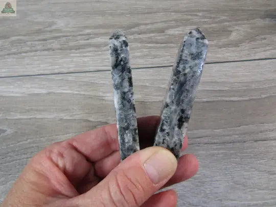 A hand holds two polished Keystone Crystals Larvikite Flat Obelisks—shimmering grounding towers from Pink Platypus Emporium—standing upright on a wooden surface.