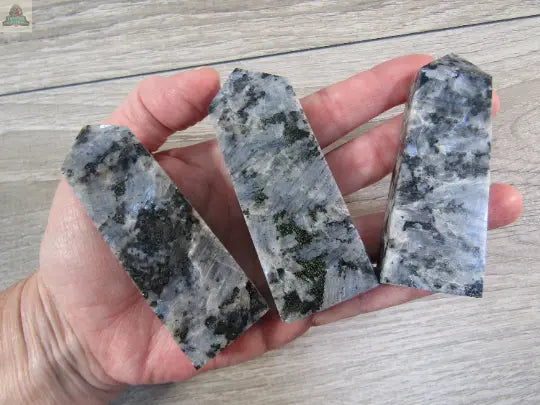 A hand displays three polished Keystone Crystals Larvikite Flat Obelisks—shimmering gray-black grounding towers from Pink Platypus Emporium—on a light wood surface, perfect for energy work.