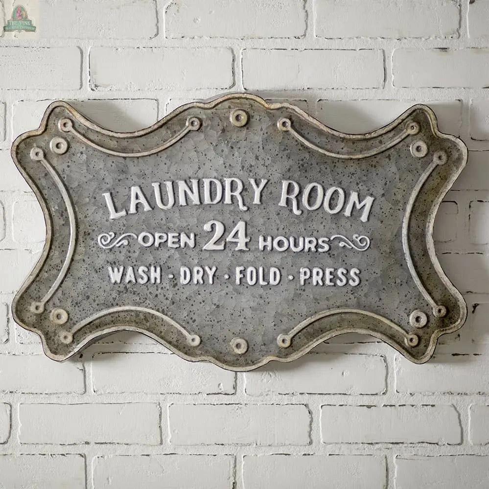 The Laundry Room Metal Sign with keyhole hangers features “Laundry Room. Open 24 hours. Wash • Dry • Fold • Press” in white lettering with ornate edging, creating charming wall decor on a white brick wall.