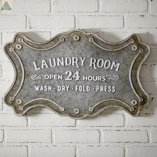 The Laundry Room Metal Sign with keyhole hangers features “Laundry Room. Open 24 hours. Wash • Dry • Fold • Press” in white lettering with ornate edging, creating charming wall decor on a white brick wall.