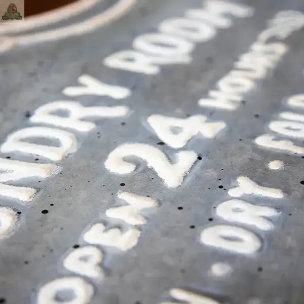 Close-up of the Laundry Room Metal Sign with raised white "OPEN 24" text on a grey textured surface. Keyhole hangers on the back make wall mounting simple. Other words are partially visible but blurred in the background.