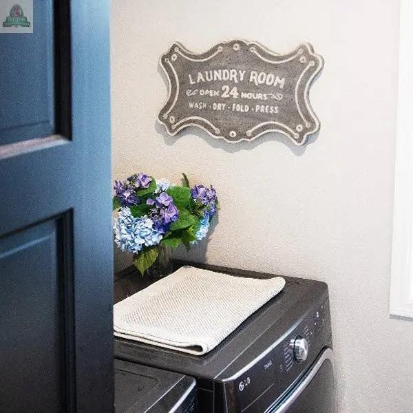 A laundry room with a washer, a folded towel, a vase of blue and purple flowers, and the Laundry Room Metal Sign featuring keyhole hangers and the words “Laundry Room, open 24 hours, wash, dry, fold, press.”.