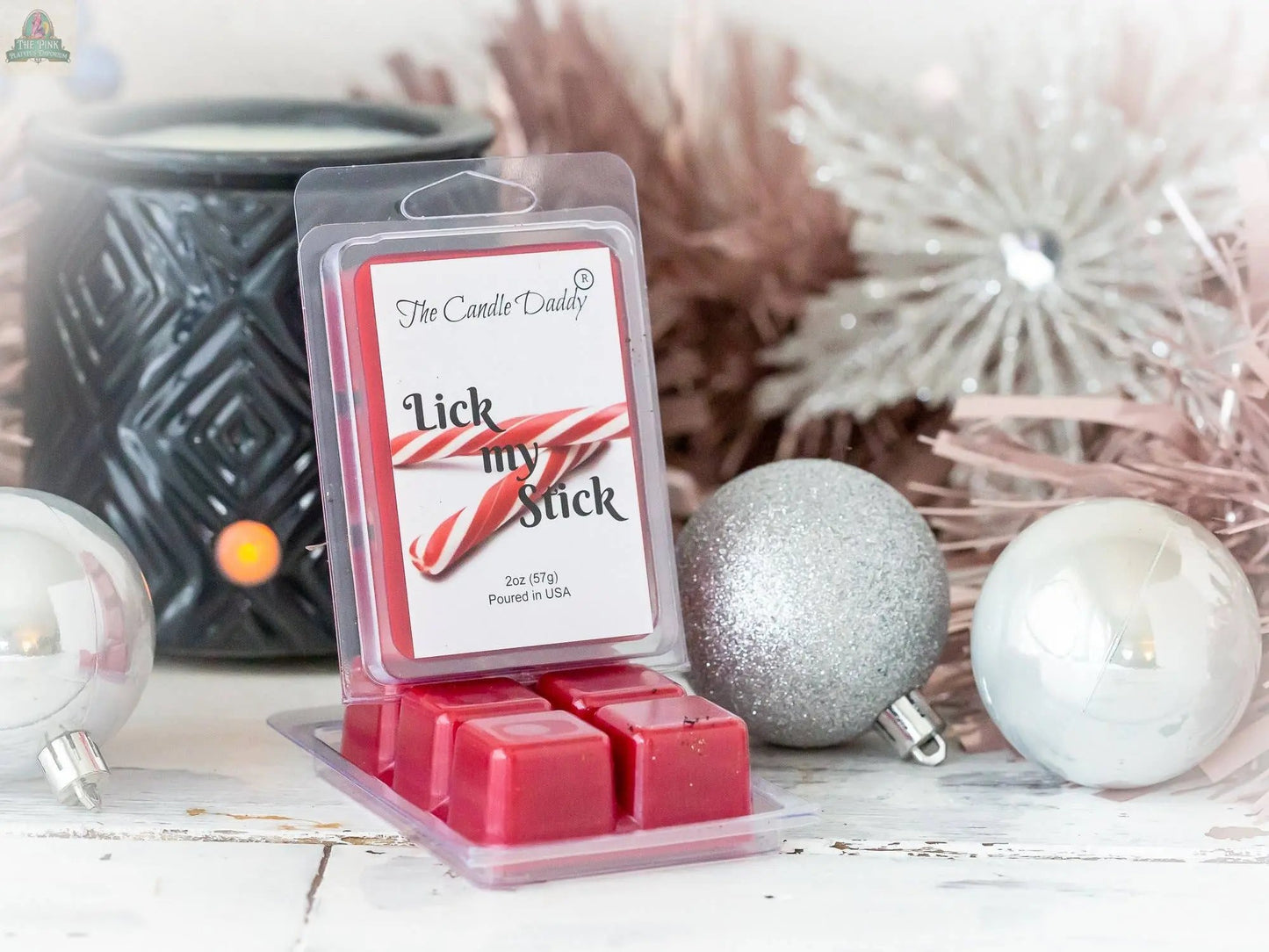 A package of Lick My Stick - Funny - Peppermint Christmas wax melts is shown among silver and white ornaments, with a black wax warmer and festive greenery in the background.