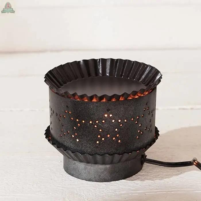 The Electric Tart Warmer-Pewter features a black metal, cupcake-liner design with a pewter finish. Star-shaped holes emit a soft glow while it sits elegantly on a white surface.