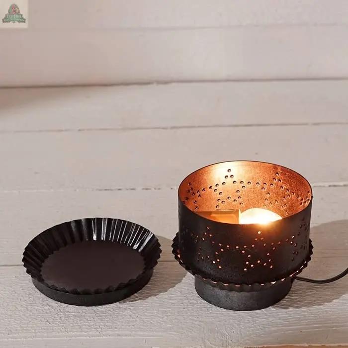 The Electric Tart Warmer-Pewter features a sleek black metal exterior and perforated copper interior, sitting on a white wooden surface beside an empty fluted black tray. It warms wax electrically, casting a cozy glow without the need for an open flame.