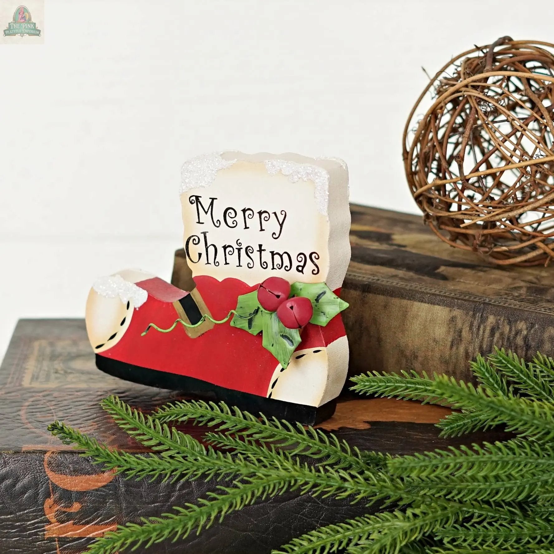 The Impressive Enterprises, Inc. Wood Red/White Snow Boot sign (5" x 4.25") adds a festive touch, featuring holly and a Merry Christmas greeting—perfect for holiday décor alongside books, twig balls, or pine branches.