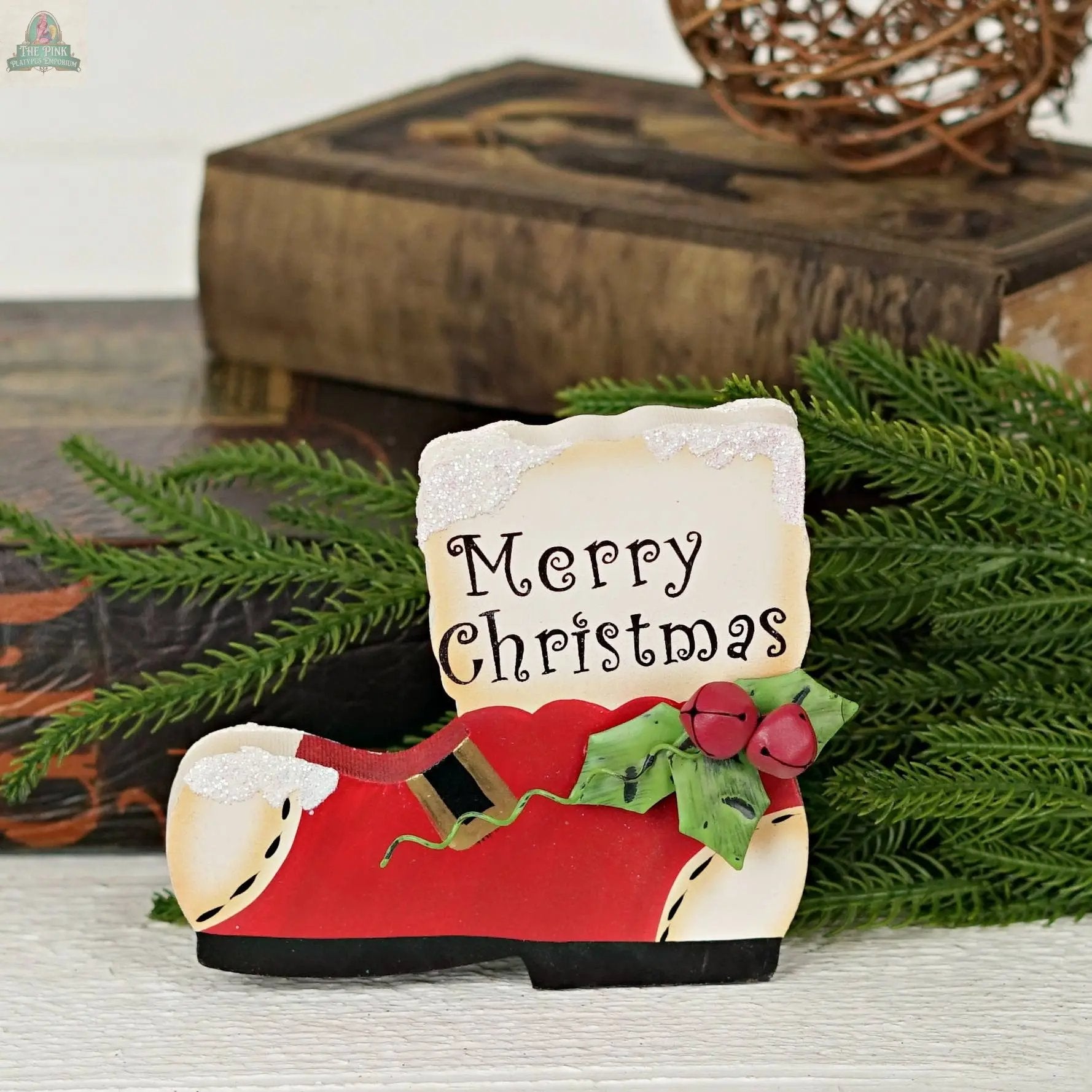 The Impressive Enterprises, Inc. Wood Red/White Snow Boot Sign (5" x 4.25") features holly leaves and berries with a "Merry Christmas" sign, displayed among evergreen branches and old books for a festive touch.