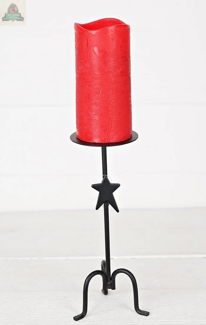 A red pillar candle rests on the Forged Iron Candle Holder, which features a star accent on its stem, creating a striking tabletop display on a light wooden surface.