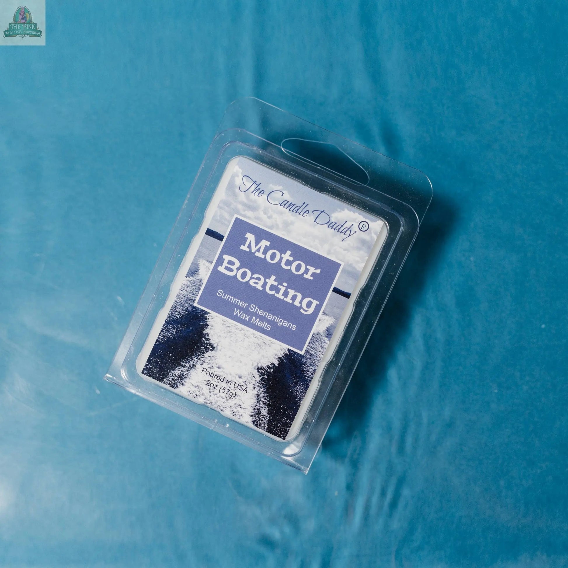 A 2oz pack of MOTOR BOATING - SUMMER SHENANIGANS SCENTED WAX MELT by The Candle Daddy, featuring a boat wake on the packaging, sits on a shiny blue surface—ideal for stress relief and summer relaxation.