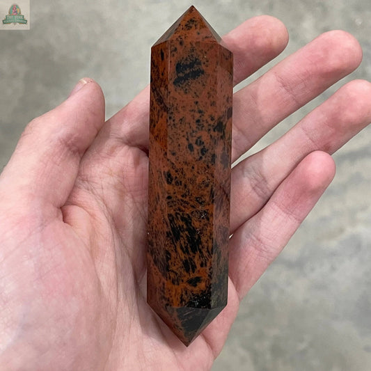 A hand holds the Keystone Crystals Mahogany Obsidian Double Terminated 3 1/2" Obelisk, showcasing its reddish-brown and black grounding patterns against a blurred gray background.