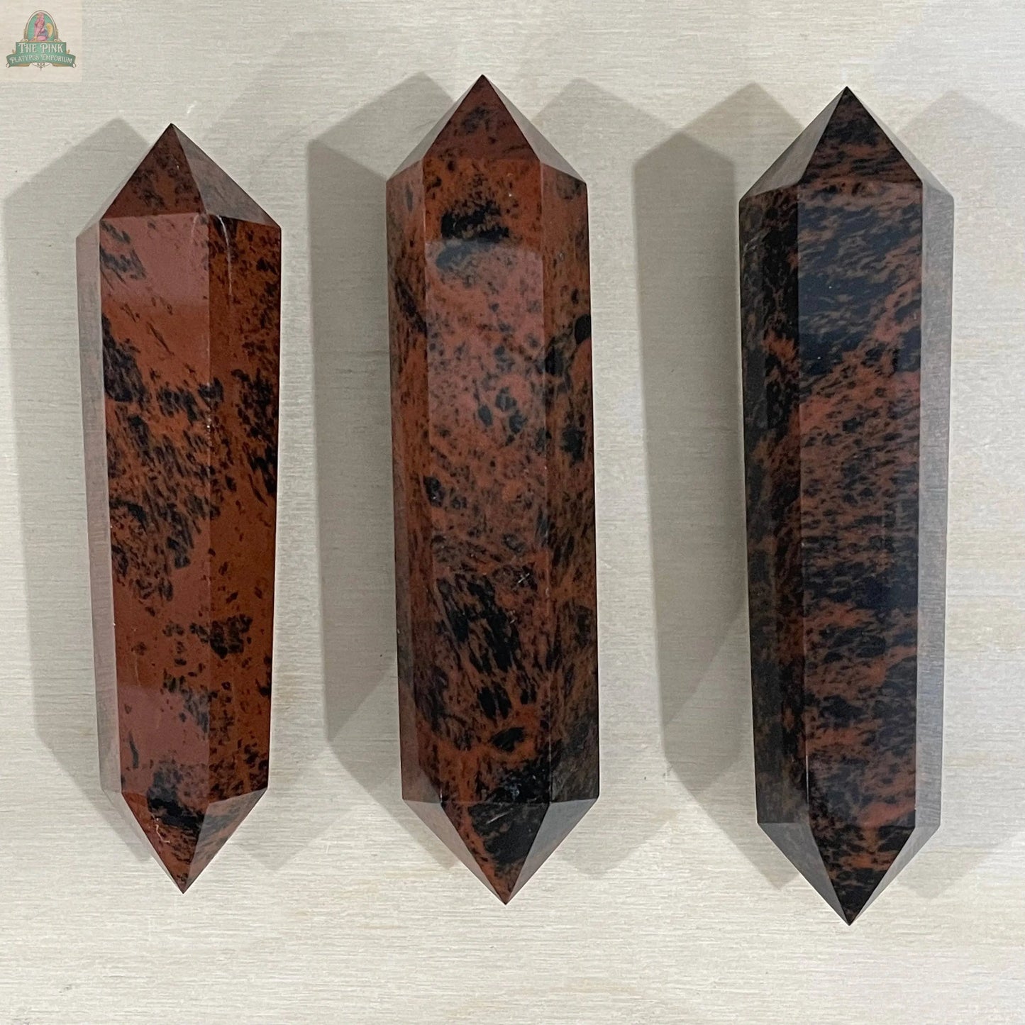 Three Keystone Crystals Mahogany Obsidian Double Terminated 3 1/2" Obelisks, showcasing brown and black mottled patterns and hexagonal pointed shapes, are arranged upright on a light surface.