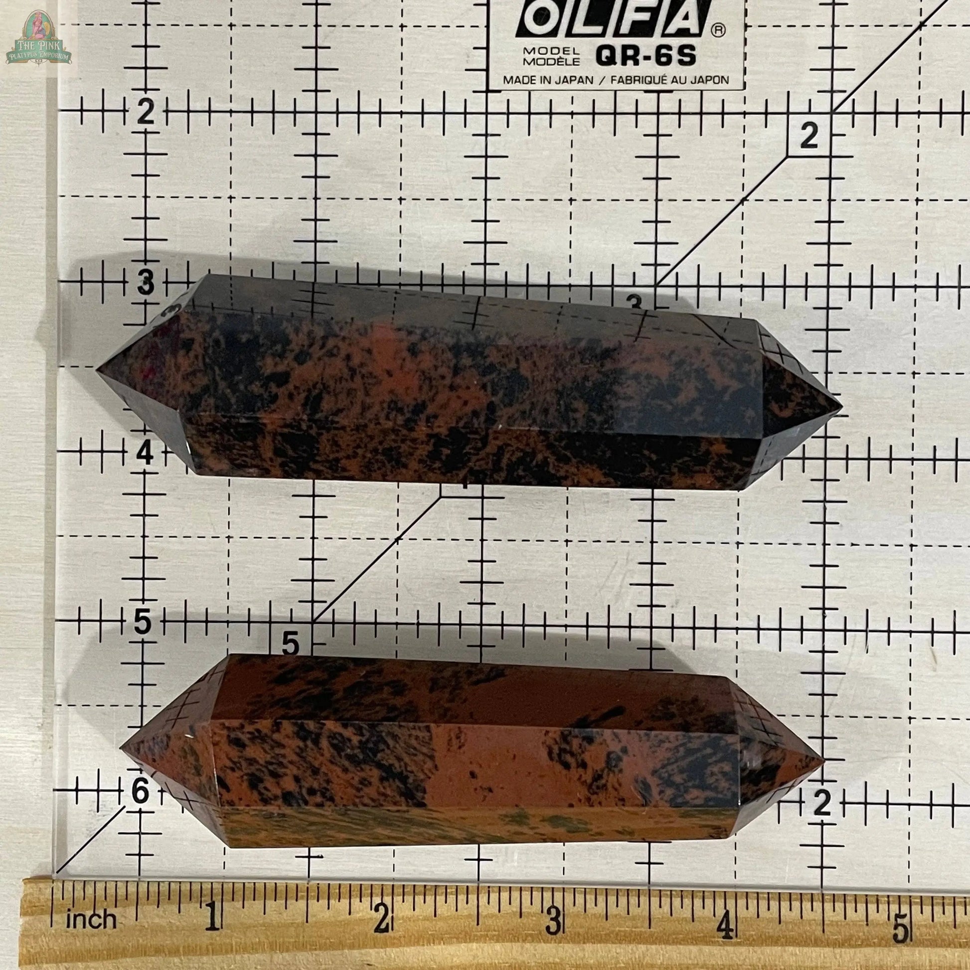 Two Keystone Crystals Mahogany Obsidian Double Terminated 3 1/2" Obelisks, featuring reddish-brown and black tones, are displayed horizontally on a cutting mat grid with ruler for scale.