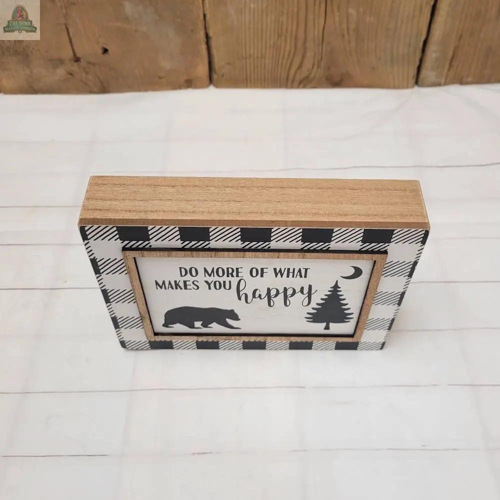The Makes You Happy Sign is a rustic wooden wall hanging with a black and white plaid border, featuring “Do more of what makes you happy” and bear, tree, and moon silhouettes—ideal for charming home decor.