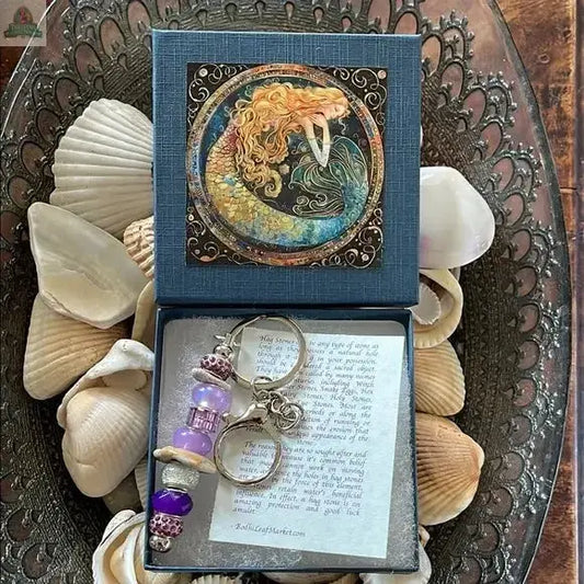 The Mermaid Keychain Gift Set features a beaded charm with purple and silver accents, a local hand chosen beach stone, and assorted Sanibel Island seashells, all beautifully arranged in a decorative box with a mermaid illustration and a tucked-in note.