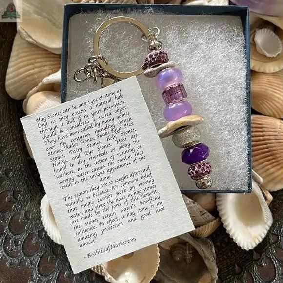 The Mermaid Keychain Gift Set features purple and silver beads, shells, and a locally chosen beach stone from Sanibel Island in a gift box. Includes a card about hag stone folklore and scattered seashells for decoration.