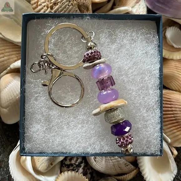 The Mermaid Keychain Gift Set features a charm with purple, silver, and beige beads, seashell pieces, and a hand-chosen Sanibel Island beach stone. It comes in a white-cushioned gift box, surrounded by seashells reminiscent of coastal treasures.