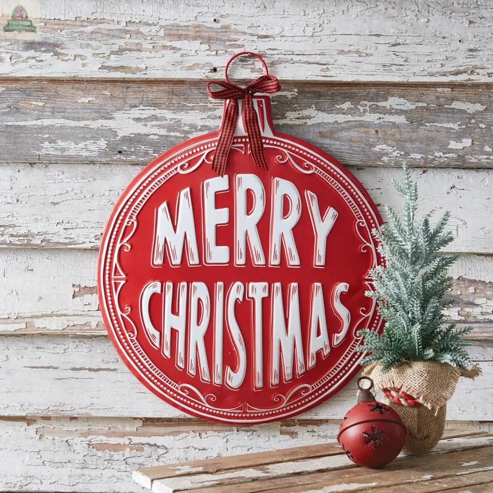 The Merry Christmas Ornament Wall Decor, featuring bold white letters, hangs on a rustic wooden wall beside a potted pine tree and red bauble, adding festive charm to your holiday decorations.