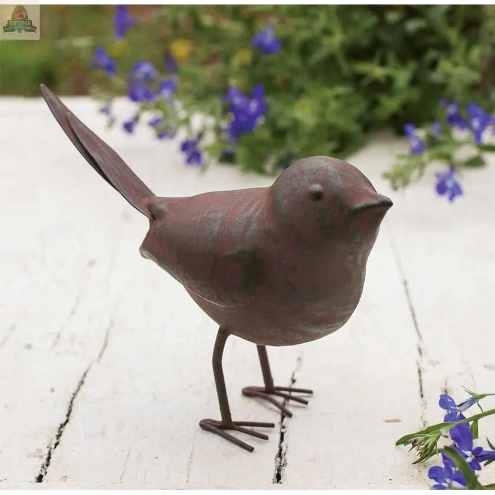 The Metal Songbird, featuring a raised tail, stands on a weathered white wooden surface, creating a charming shelf accent amid green foliage and small purple flowers.