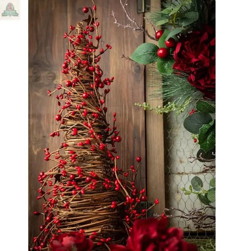The Mixed Berry Cone Tree With Rust Stars, 17"H x 5.25" base, stands before a rustic wood backdrop with green leaves and red flowers nearby, adding a festive accent with woven twigs and red berries.