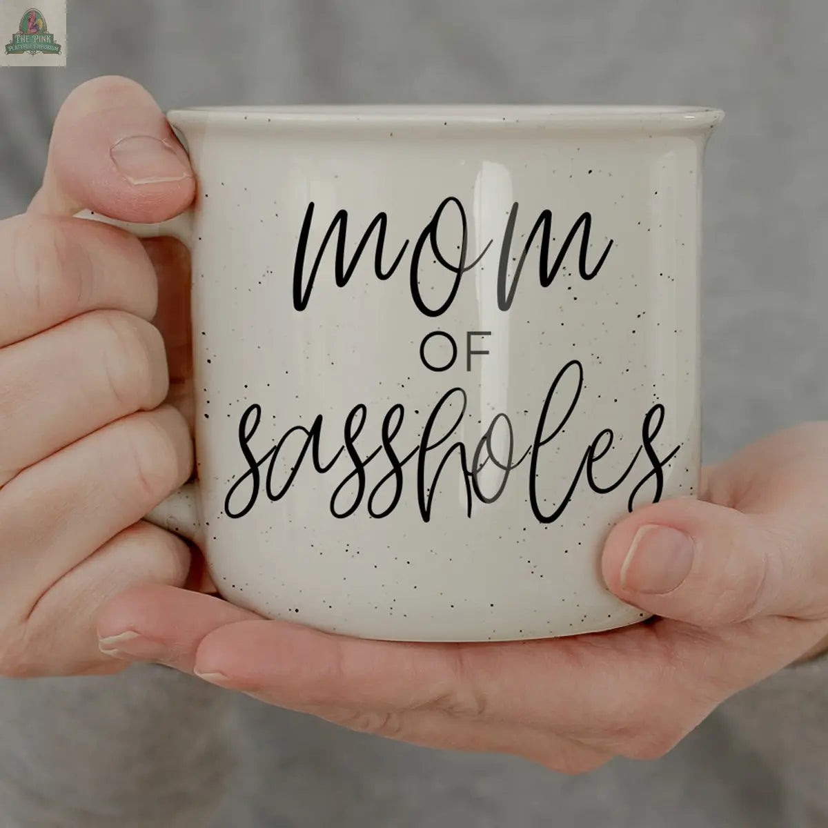 A person holds a 14.5oz white "Mom of Sassholes" mug—funny black cursive and print text make it the perfect gift for the ultimate Chief Sass Officer.