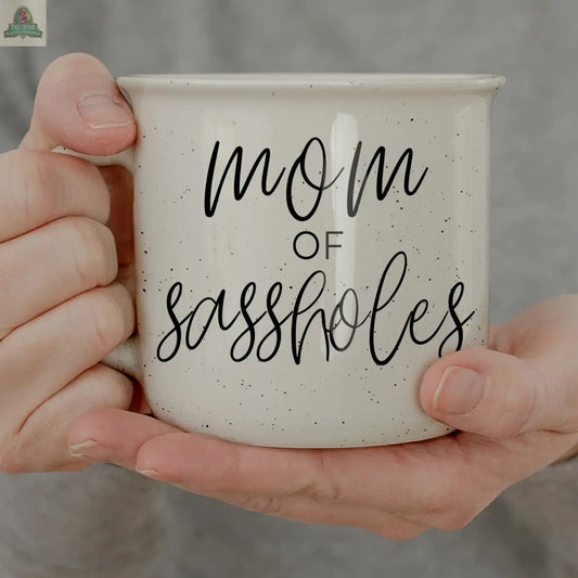 A person holds a 14.5oz white "Mom of Sassholes" mug—funny black cursive and print text make it the perfect gift for the ultimate Chief Sass Officer.