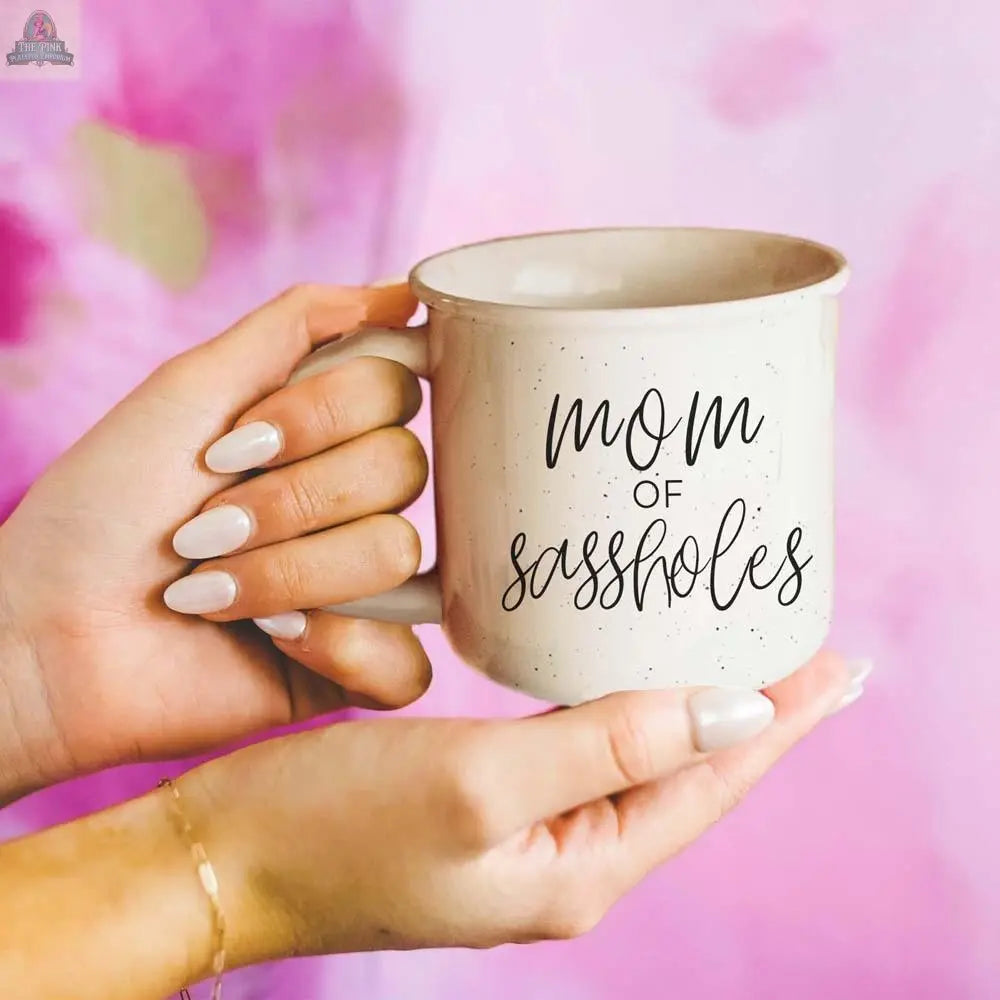 A person with manicured nails holds a beige "Mom of Sassholes" 14.5oz mug featuring black script, against a pink and purple blurred background.