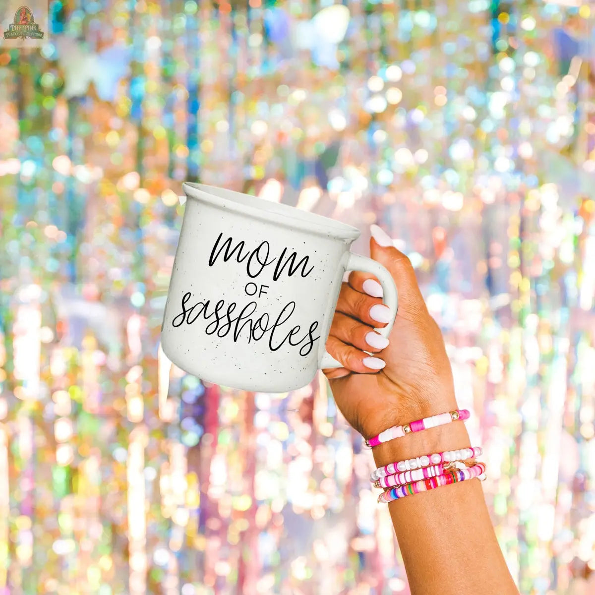 A hand with colorful beaded bracelets holds the Mom of Sassholes 14.5oz mug, labeled Chief Sass Officer, against a sparkly and colorful blurred background.