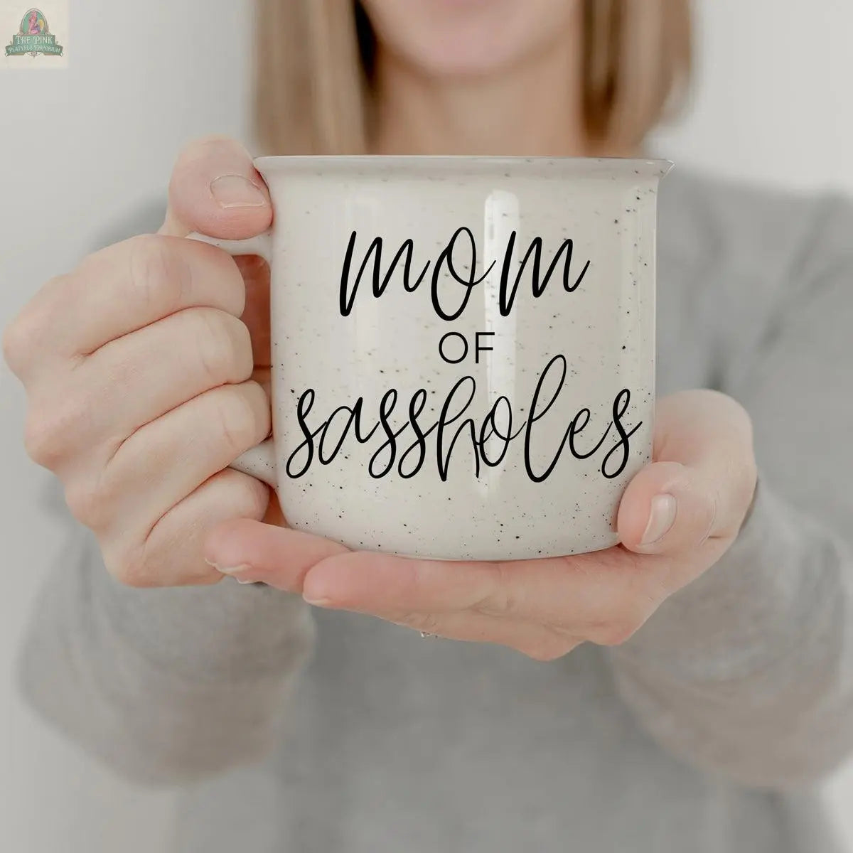 A person in a gray shirt holds the Mom of Sassholes 14.5oz white speckled mug with black cursive lettering—a funny parent mug perfect for mastering sass management.