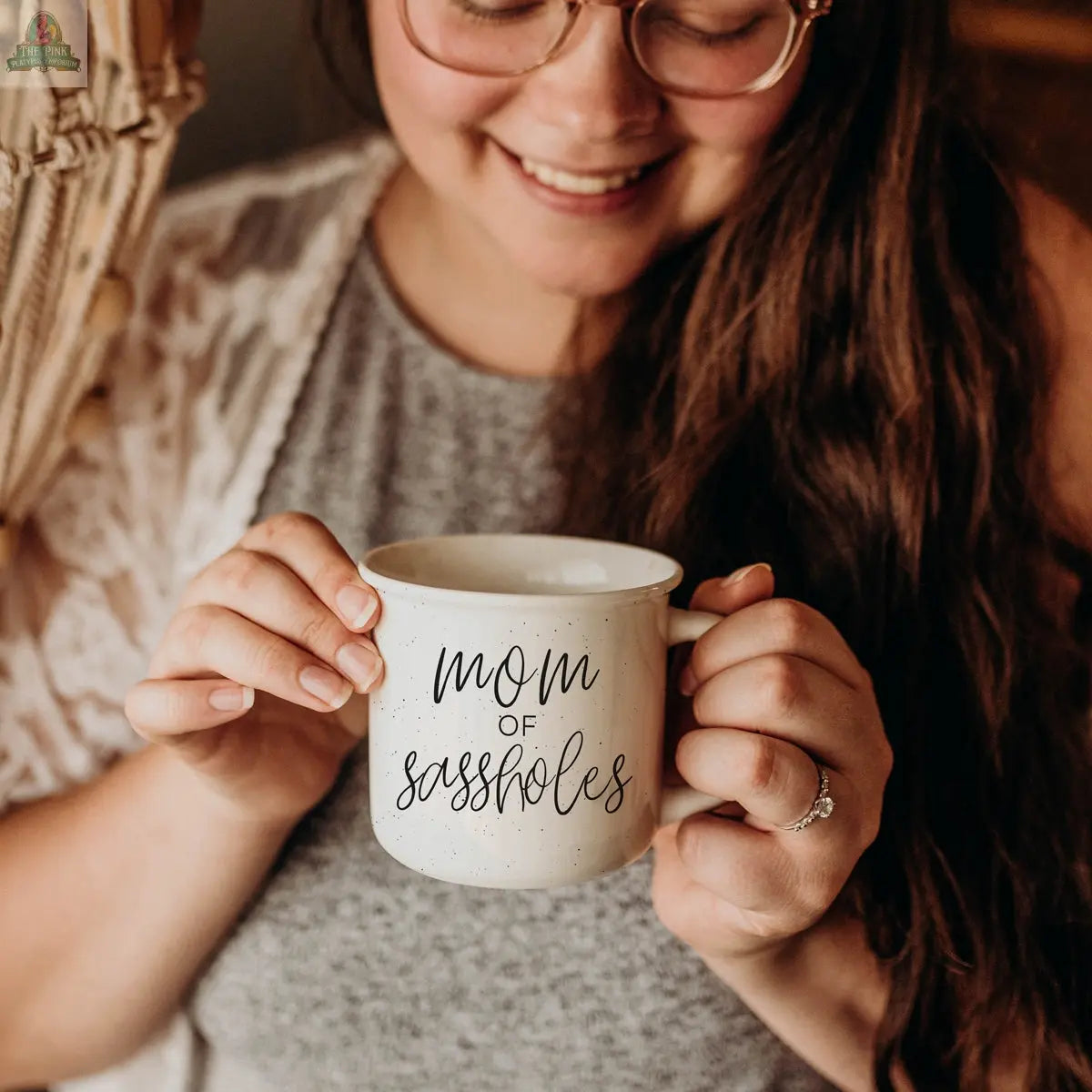 A smiling woman with long brown hair and glasses holds the “Mom of Sassholes 14.5oz” mug. She wears a gray top and displays an engagement ring on her finger.