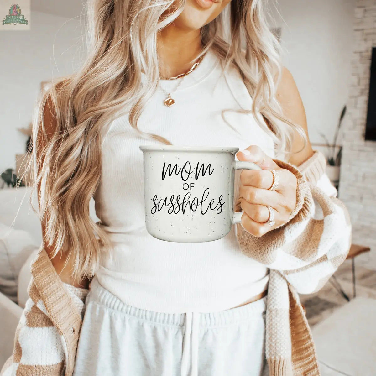 A woman with long blonde hair, in a cream tank top and cozy cardigan, holds the Mom of Sassholes 14.5oz mug featuring black script. She stands in a stylish, modern living room.
