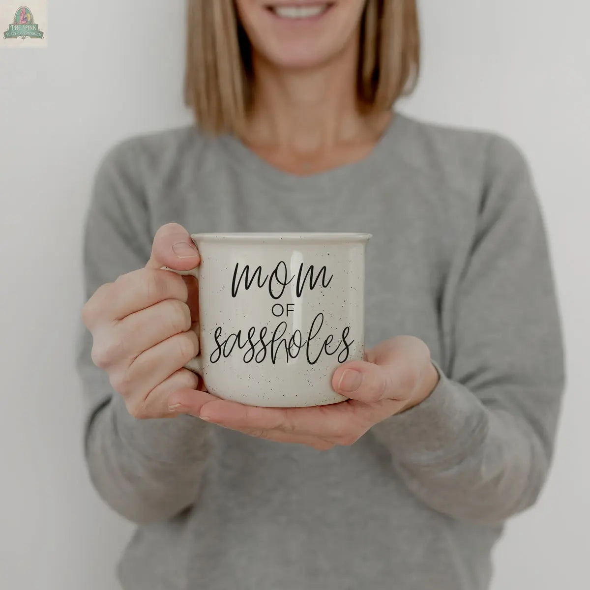 A woman in a gray sweater holds the "Mom of Sassholes 14.5oz" white mug with cursive text—an ideal funny parent gift. The background is plain and light-colored.