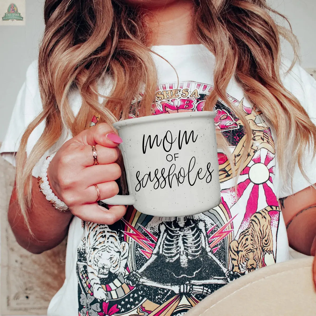 A woman with long wavy hair holds the "Mom of Sassholes 14.5oz" mug near her chest, wearing a colorful graphic tee with tigers, stars, and rainbows. Only her torso is shown.