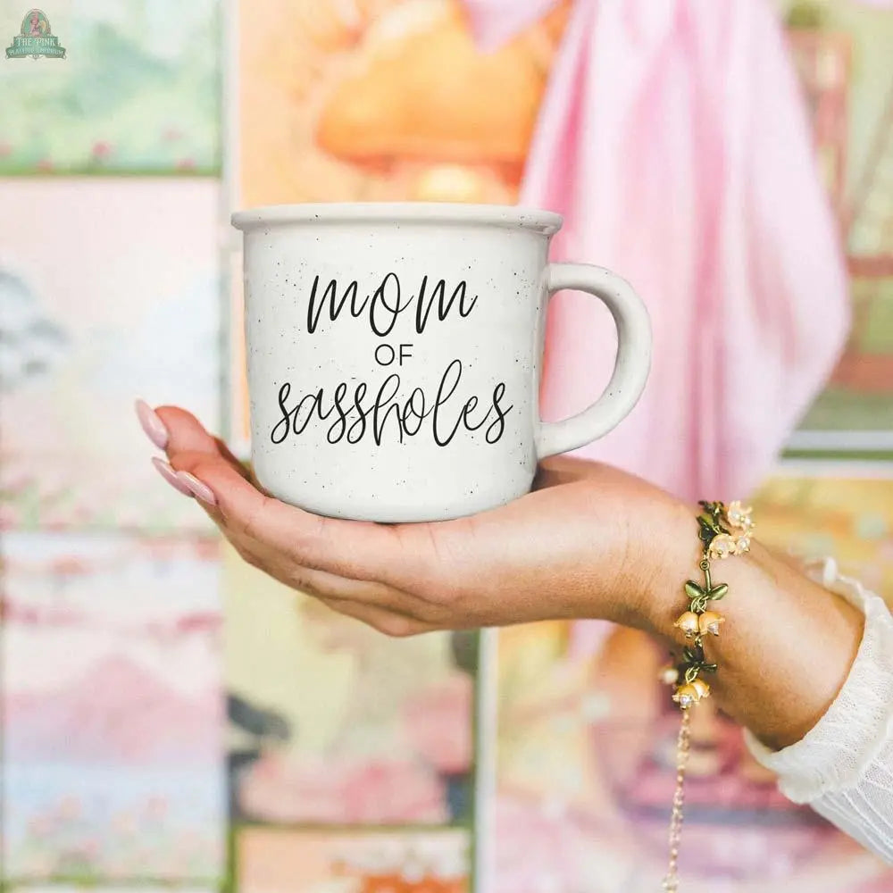 A hand wearing a beaded bracelet holds the Mom of Sassholes 14.5oz mug, featuring "mom of sassholes" in black script, with a colorful, blurred background.