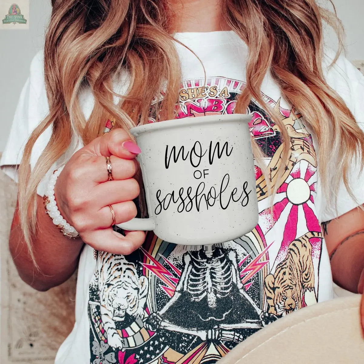 A woman with wavy hair holds a "Mom of Sassholes 14.5oz" mug. She wears a colorful graphic tee with tigers, skeletons, and text. Only her torso and hand are visible.