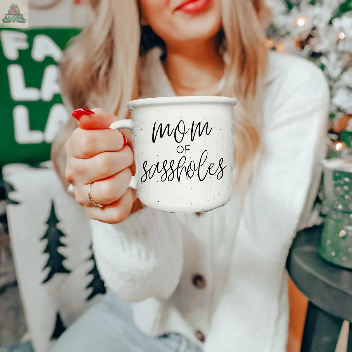 A smiling woman with long blonde hair holds the "Mom of Sassholes 14.5oz" mug. She wears a light cardigan and sits near holiday decor, including a green pillow and Christmas tree. The mug features black script text.