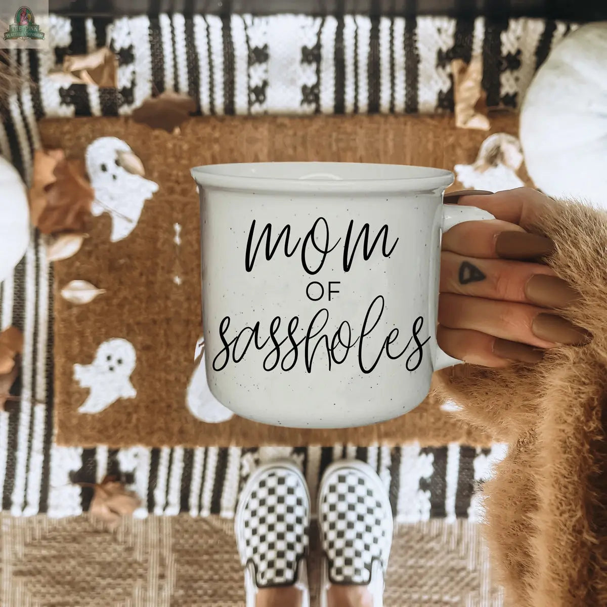 A person with long nails holds a Mom of Sassholes 14.5oz mug. Below are black-and-white checkered slip-on shoes, a doormat with white ghosts, and scattered autumn leaves.