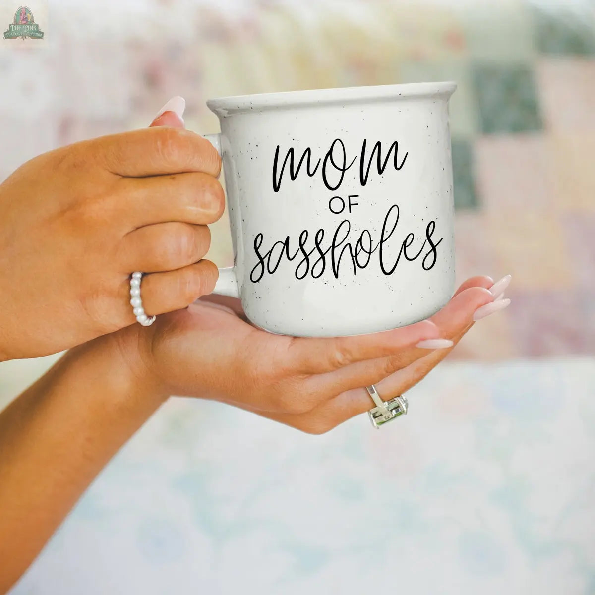A person with manicured nails and rings holds the Mom of Sassholes 14.5oz mug featuring black script, with a softly blurred pastel background adding a playful vibe to this cheeky parent gift display.