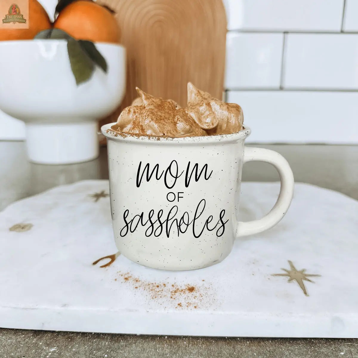 A beige speckled Mom of Sassholes 14.5oz mug with “Chief Sass Officer” sits on a marble board, filled with a frothy cinnamon-topped drink. A white bowl of oranges and a wooden cutting board are in the background.