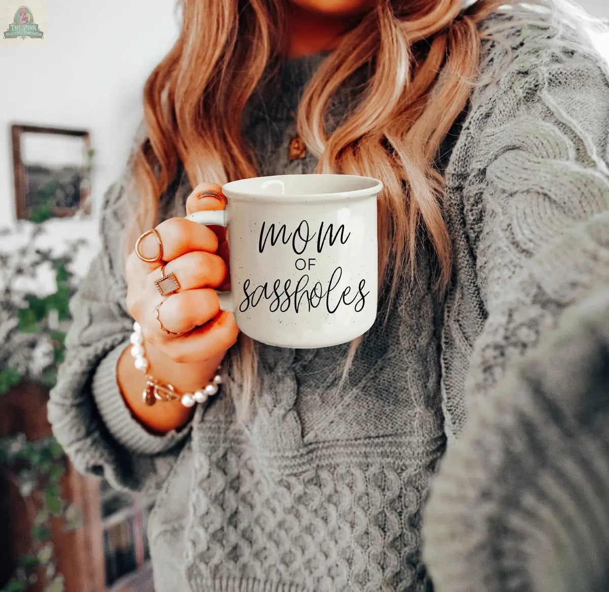 A woman in a gray sweater holds the “Mom of Sassholes 14.5oz” mug in black script, accessorized with rings and a bracelet. Her long hair is loose, and a softly blurred background draws focus to this playful parent mug.