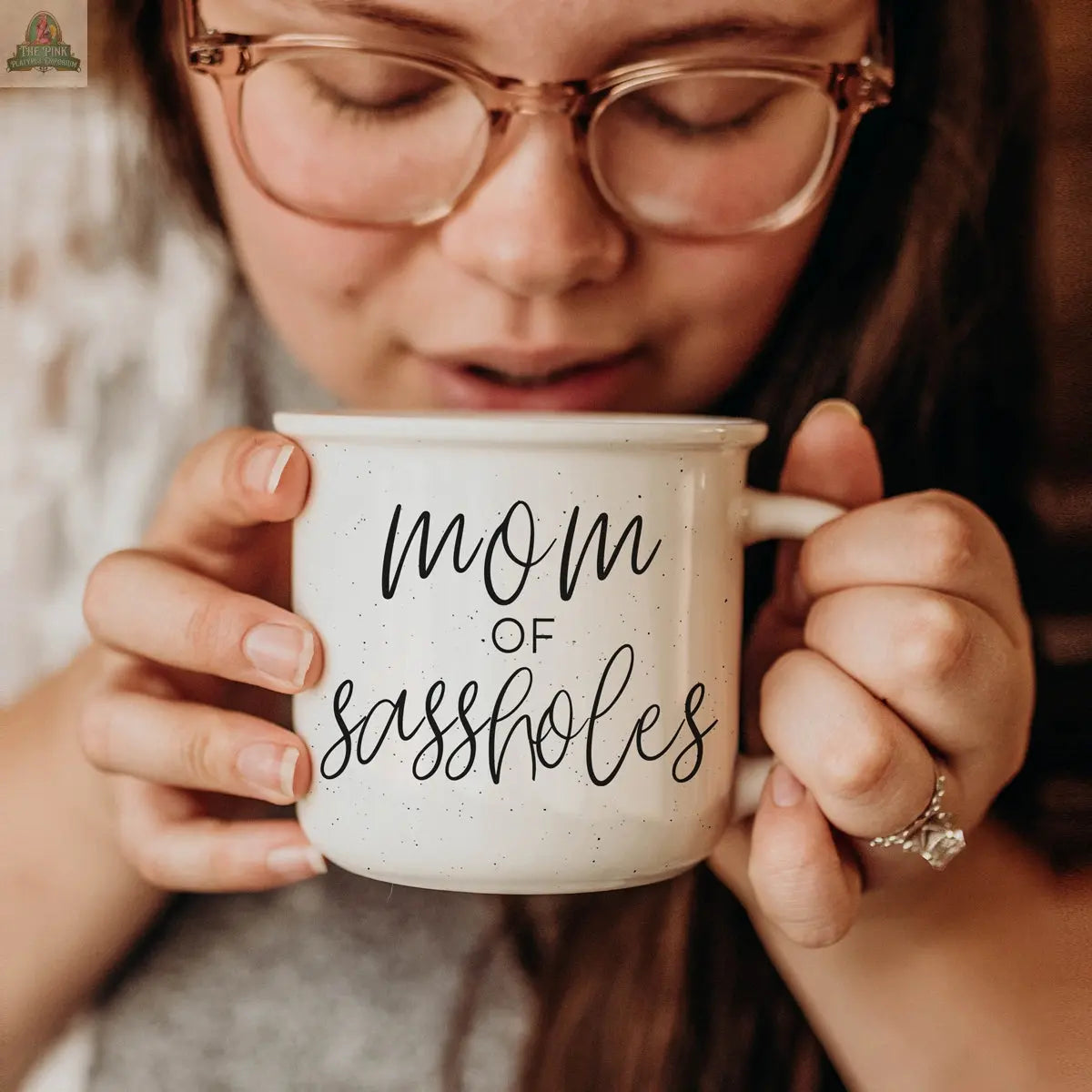 A woman with glasses holds the 14.5oz Mom of Sassholes mug near her face. The white mug features “mom of sassholes” in black script, and she wears a ring on her finger.