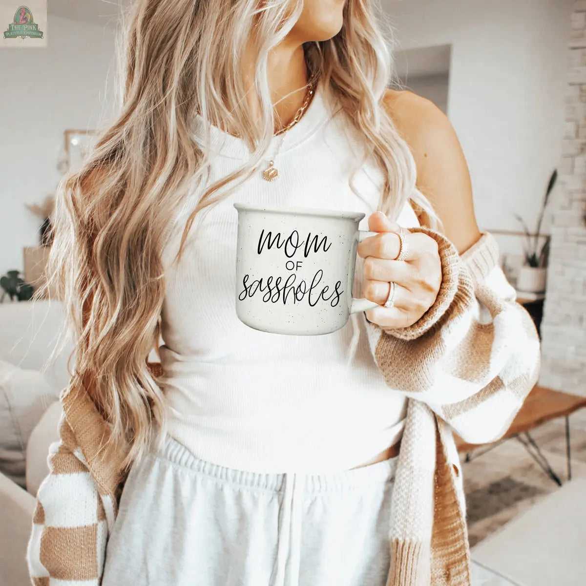 A woman with long blonde hair holds the Mom of Sassholes 14.5oz mug, wearing a white tank top, light gray sweatpants, and a brown striped cardigan in a cozy, modern living room.