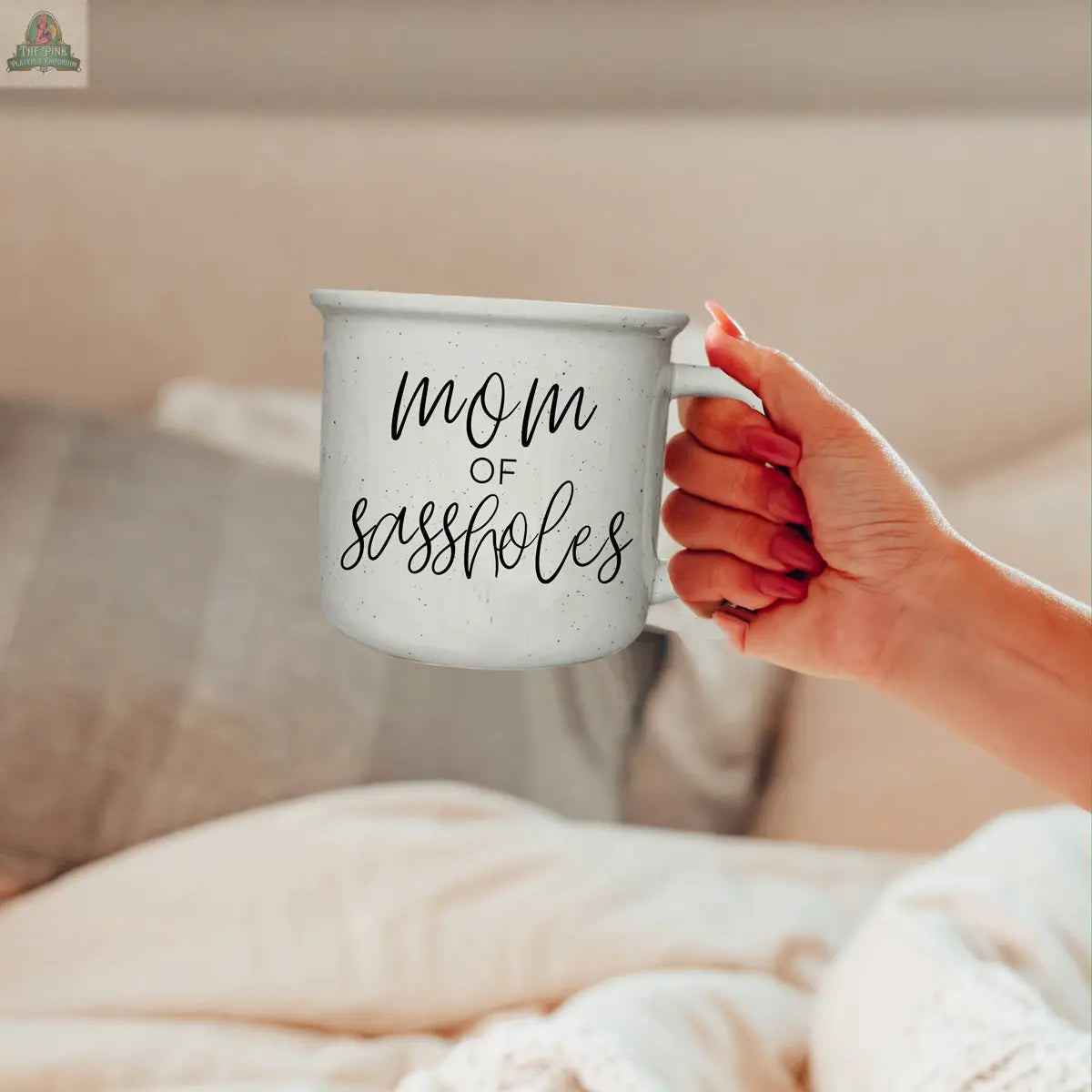 A hand holds the "Mom of Sassholes 14.5oz" white speckled mug with bold cursive text. The scene is cozy, featuring a bed with soft pillows and a blanket, perfectly complementing this playful parent mug.