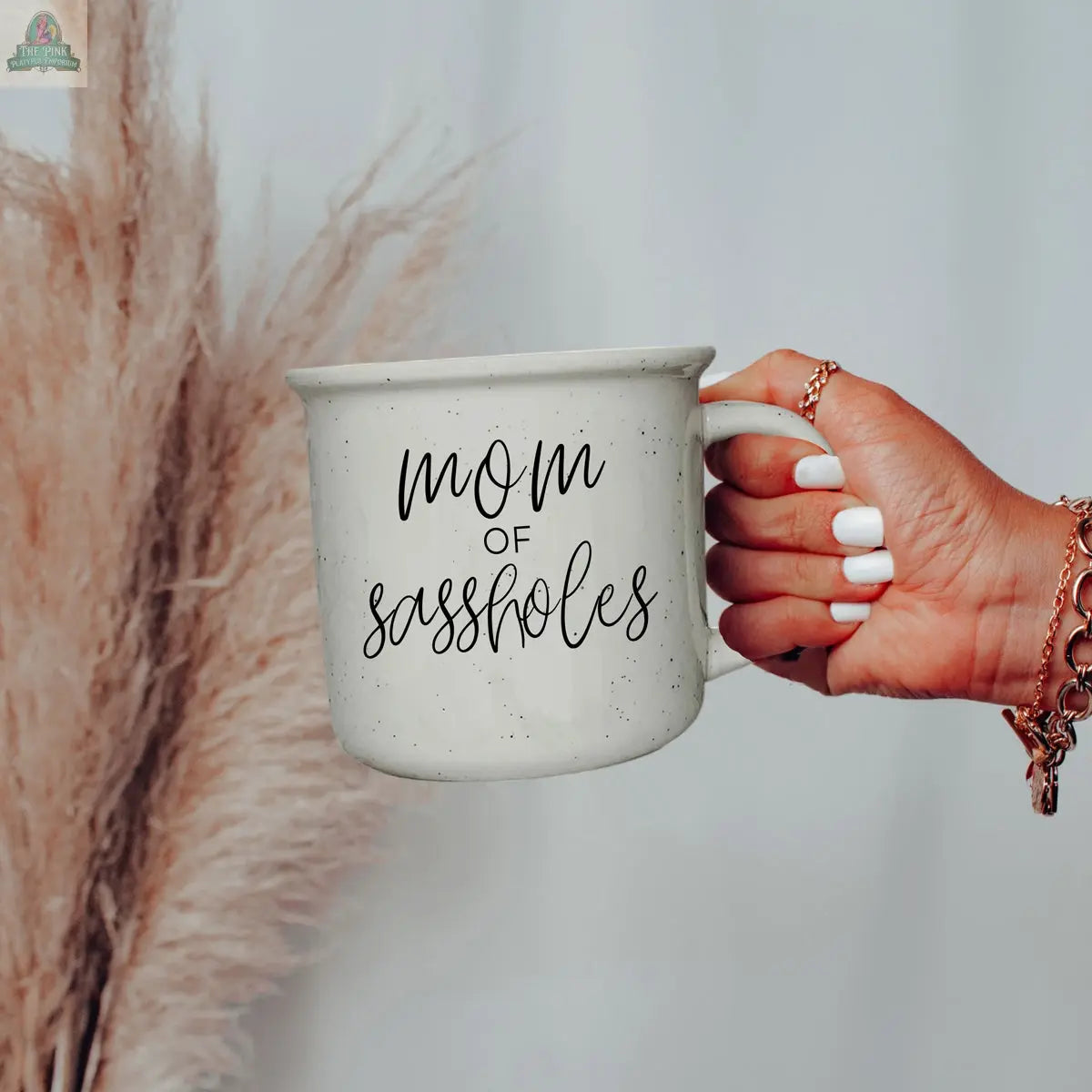 A hand with white-painted nails holds a "Mom of Sassholes 14.5oz" mug in black script, with blurred beige pampas grass in the background.