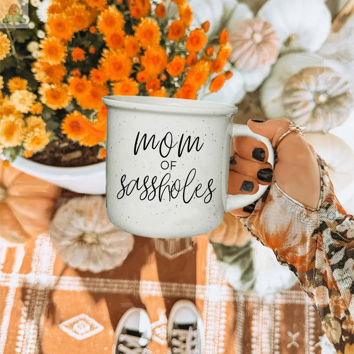 A hand with patterned sleeves holds a Mom of Sassholes 14.5oz mug; in the background are orange flowers, white pumpkins, and an orange patterned rug.