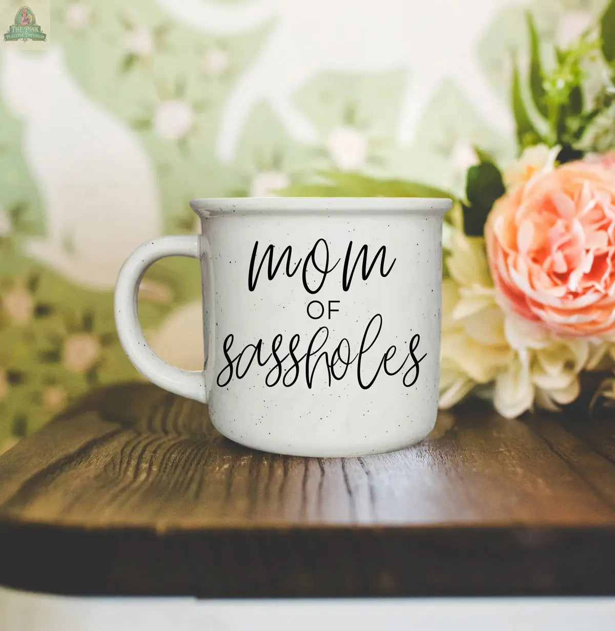 A Mom of Sassholes 14.5oz white mug with “Chief Sass Officer” and “mom of sassholes” in black script sits on a wooden surface, with blurred flowers and a green and white patterned wall enhancing this playful parent mug scene.