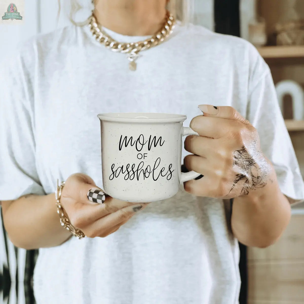 A woman with painted nails holds the "Mom of Sassholes 14.5oz" mug, featuring sassy text. She wears a white t-shirt, gold jewelry, and has black ink stains on her left hand. The indoor background is softly blurred for a warm feel.