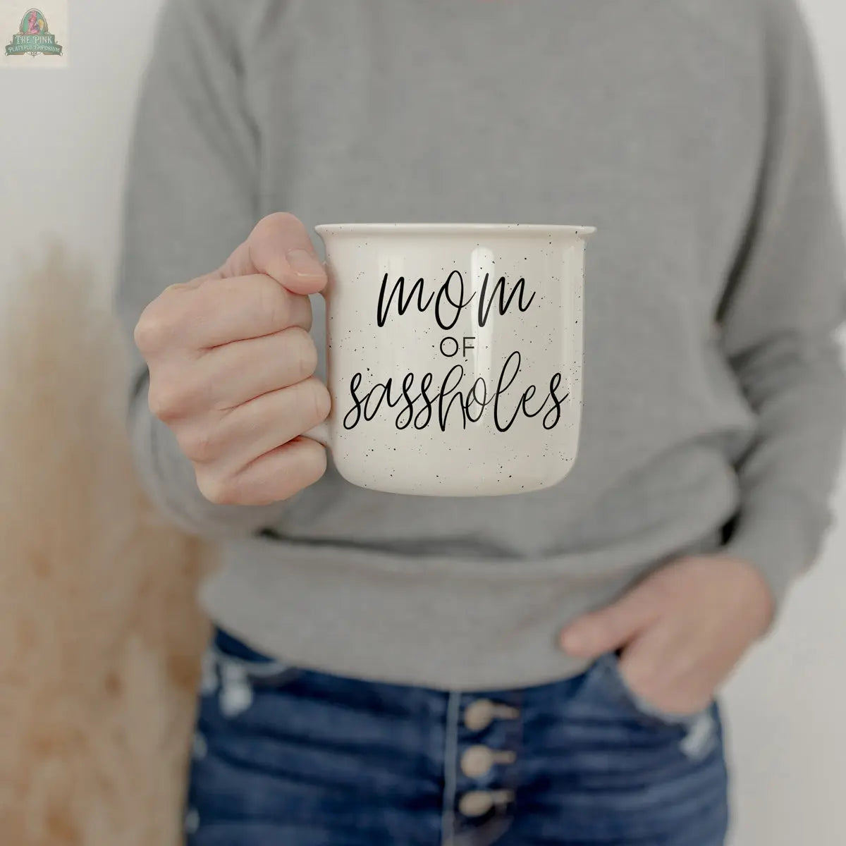 A person in a gray sweatshirt and blue jeans holds a white speckled Mom of Sassholes 14.5oz mug with the words "mom of sassholes" written on it in black script.