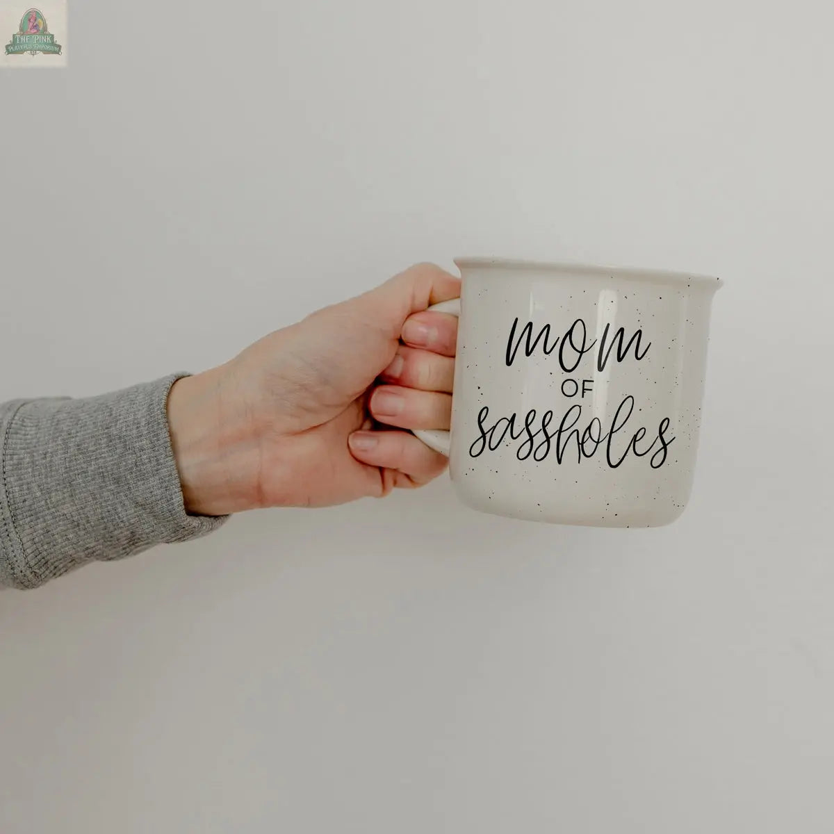 A hand holds the Mom of Sassholes 14.5oz white speckled mug, featuring "mom of sassholes" in black cursive and uppercase letters—a fun parent mug shown against a plain light background.