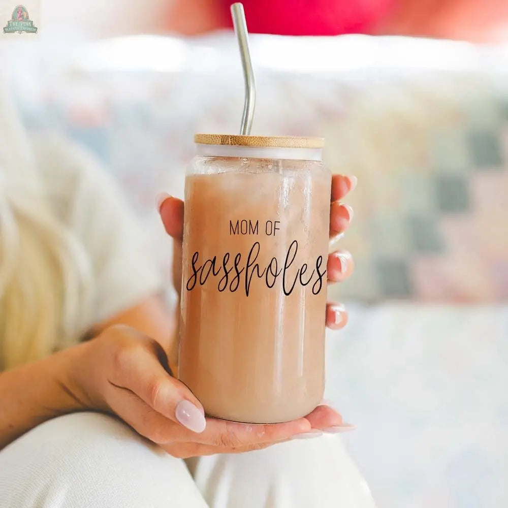 A person holds a 17oz Mom of Sassholes mason jar, topped with a bamboo lid and stainless steel straw, filled with a light brown drink. The glass features "MOM OF sassholes" in bold black script.