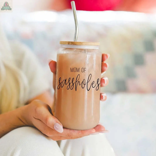 A person holds a 17oz Mom of Sassholes mason jar, topped with a bamboo lid and stainless steel straw, filled with a light brown drink. The glass features "MOM OF sassholes" in bold black script.